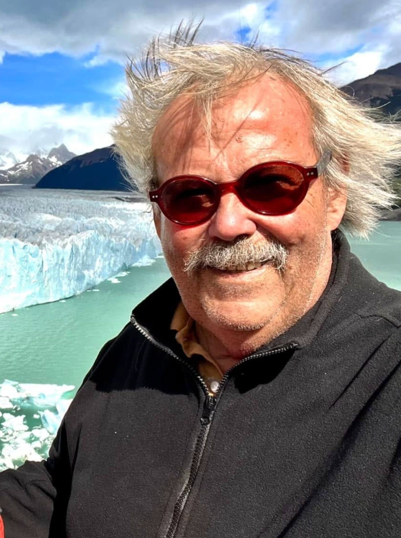 Antoine Agapitos, cheveux blancs ébouriffés et lunettes de soleil, devant un glacier et un lac turquoise.