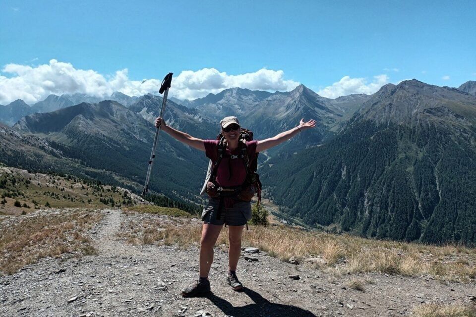 Flora Klemencie, sac à dos et bâton levé, sur un sentier de montagne face à des sommets.