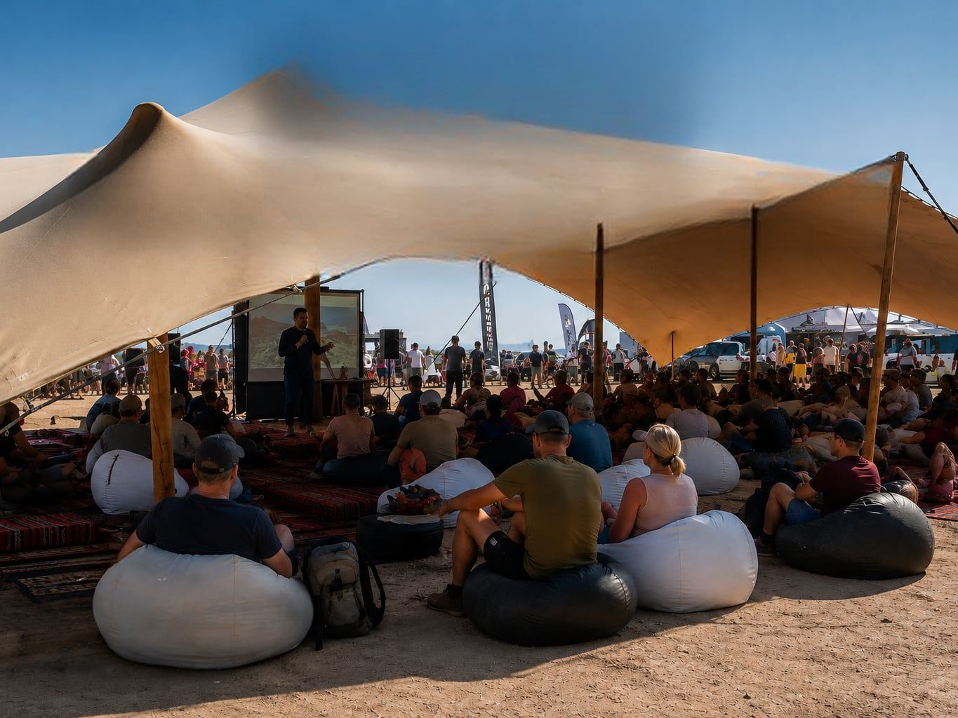 Sous une grande toile tendue, un orateur présente des images projetées devant un public assis sur des poufs et des tapis.