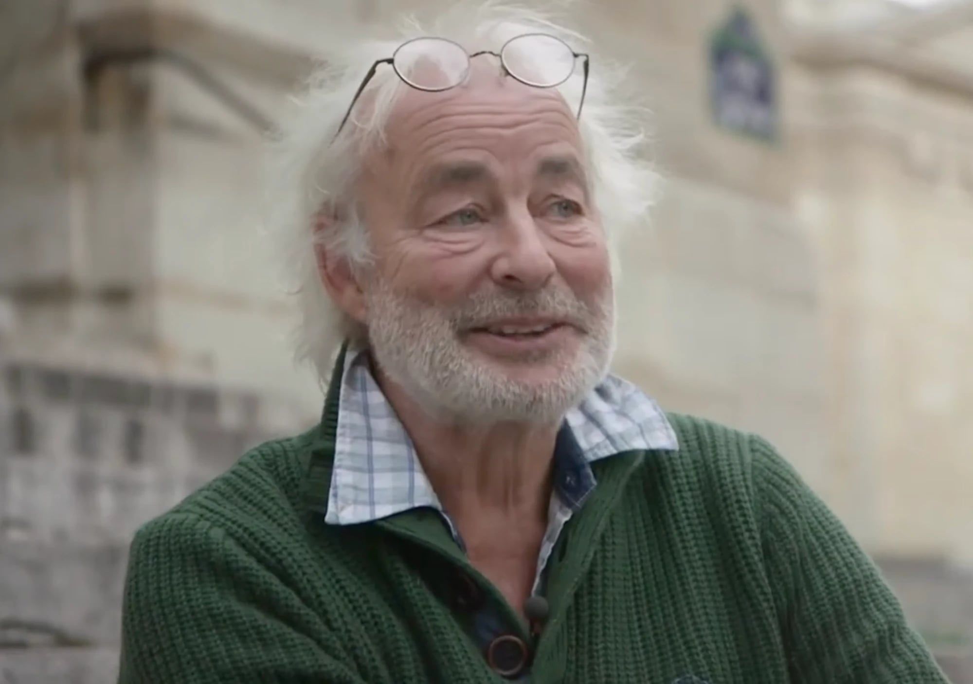 Portrait de Jack Bollet, cheveux blancs et lunettes relevées sur le front, en pull vert, souriant.
