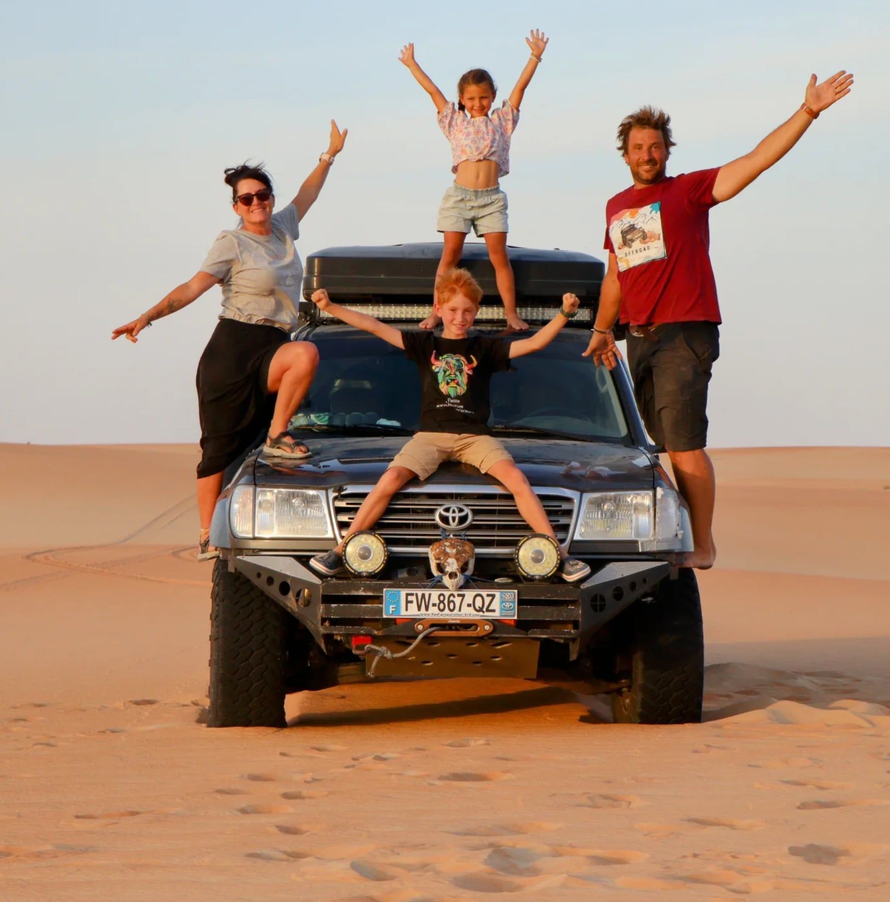 La famille Ma Tribu Trotter, bras levés, perchée sur son 4×4 au milieu des dunes de sable.