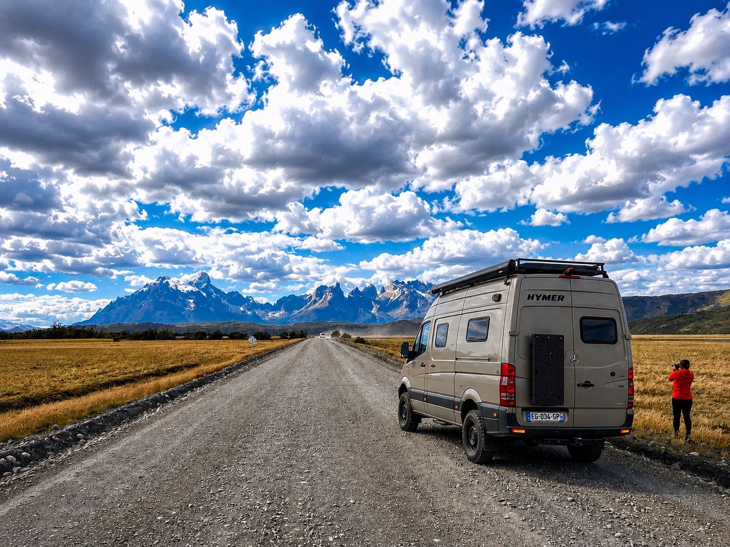 Un fourgon aménagé roule sur une piste de gravier vers des montagnes enneigées, sous un ciel chargé de nuages.