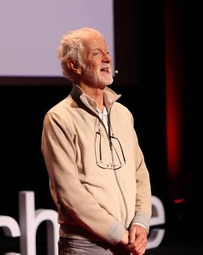 Yves Marre, cheveux et barbe blancs, en veste beige, sur scène devant un écran de conférence.